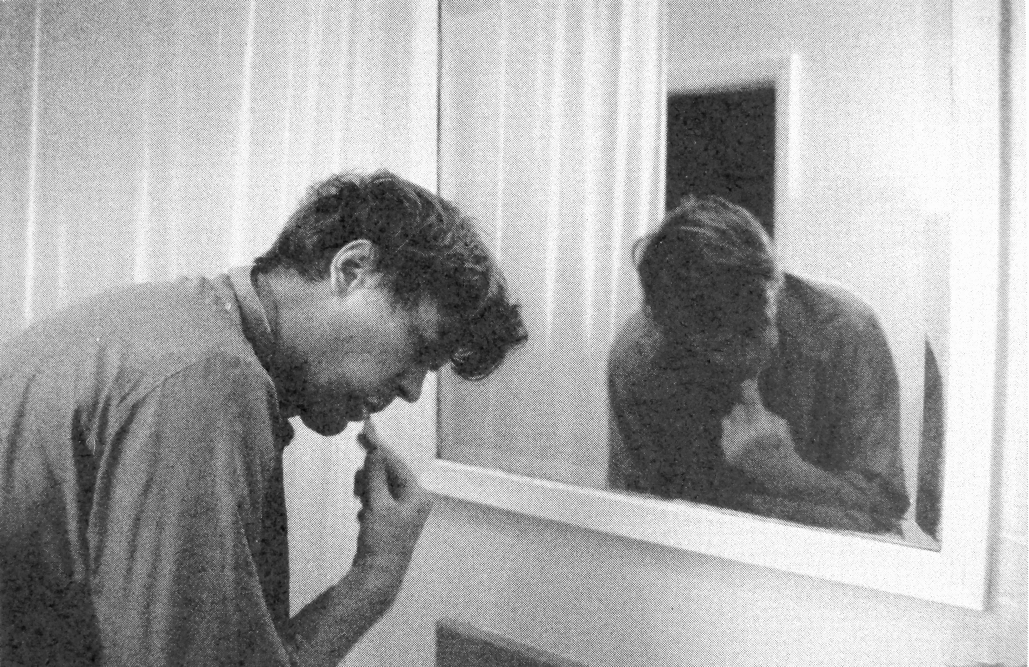 A man (Charles Allcroft)  brushes his teeth in front of a bathroom sink and mirror.